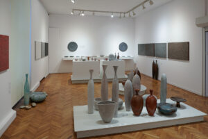 Część ekspozycji przedstawiająca prace ceramiczne wiszące na ścianach w kształcie obrazów oraz stojące w kształcie talerzy, amfor i wazonów